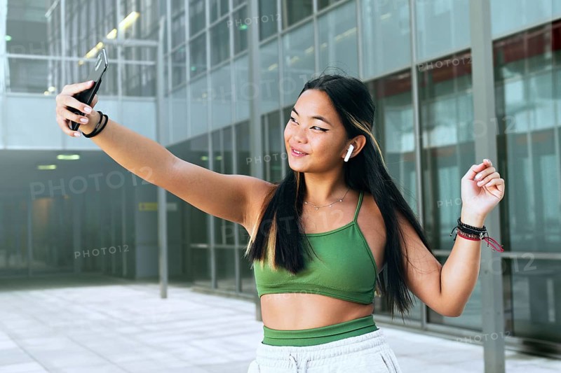 A joyful teenager takes a selfie in a modern urban setting, wearing a green top and wireless earbuds. The scene conveys a sense of youth, fun, and technology