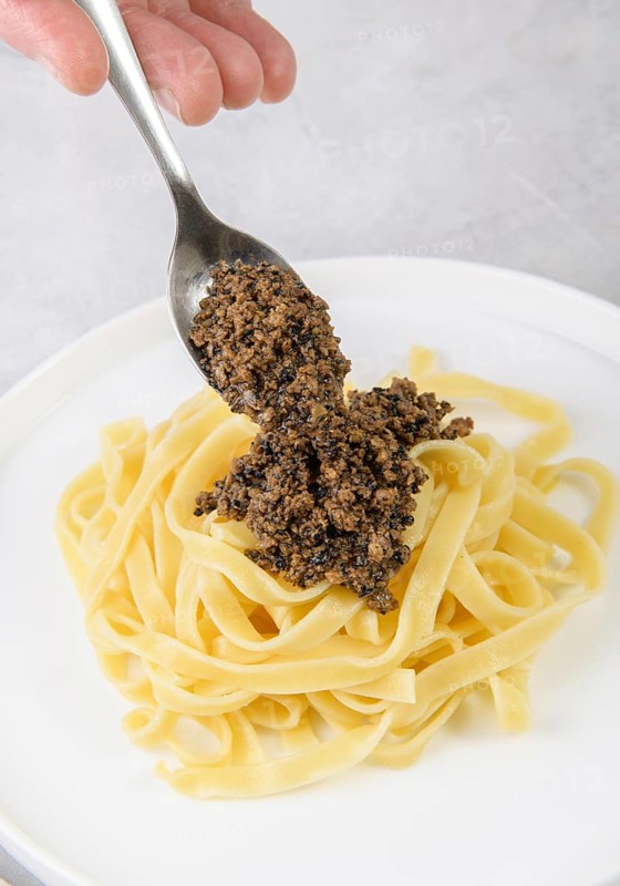 A hand elegantly spoons rich black truffle sauce onto a bed of freshly cooked tagliatelle pasta, creating a simple yet decadent Italian dish on a white plate