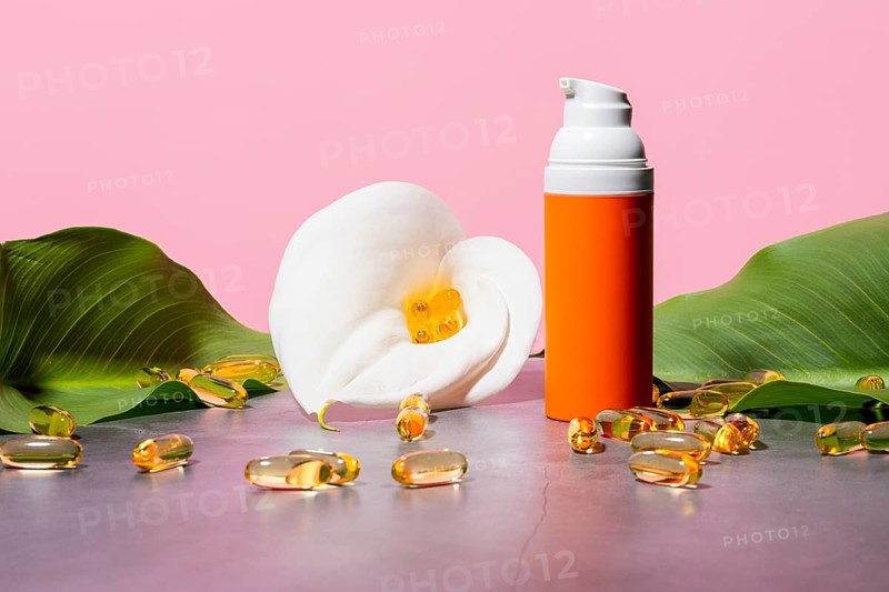 An elegant cosmetic setup featuring an orange pump bottle, surrounded by yellow omega-3 supplement capsules and a pristine white flower, accentuated by lush green leaves against a soft pink background