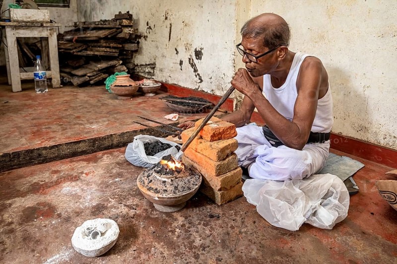 Man uses a blowpipe to spark a coal-fired vessel, a man burns sapphires in a small stove in the town of Ratnapura in Sri Lanka