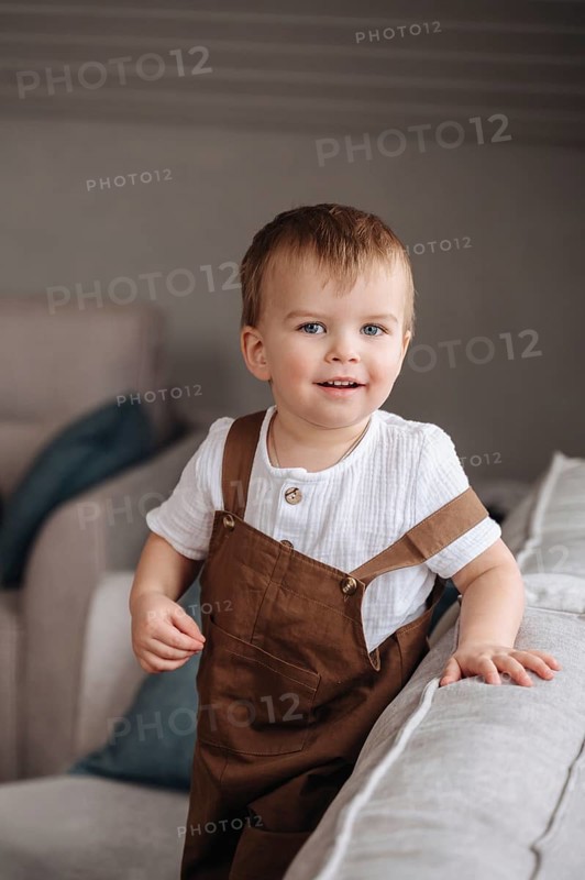 A joyful toddler smiles brightly while standing in a cozy, softly lit living room. Wearing brown overalls over a white shirt, this image captures the innocence and happiness of childhood