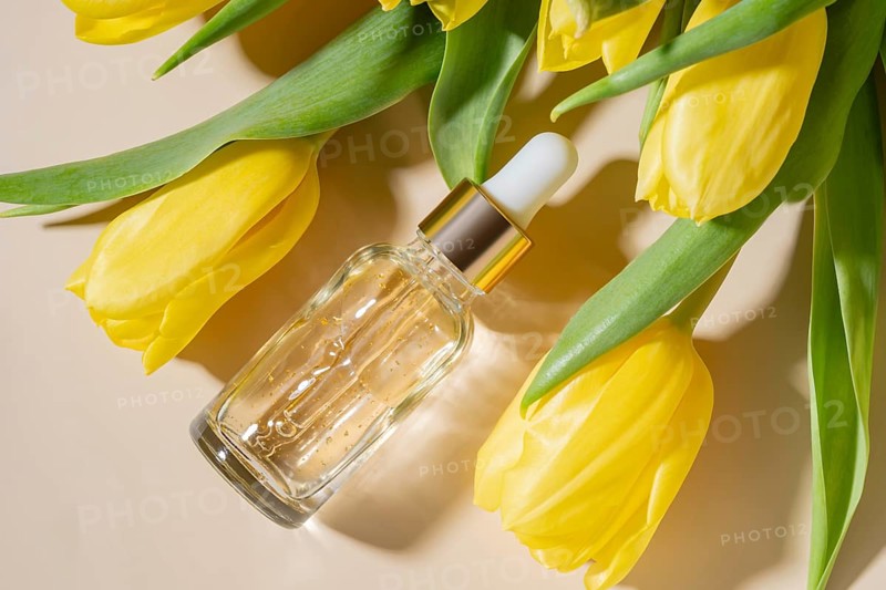 A glass dropper cosmetic of luxurious skincare serum rests among vivid yellow tulips on a soft beige background. The arrangement exudes freshness and elegance, perfect for beauty and wellness themes