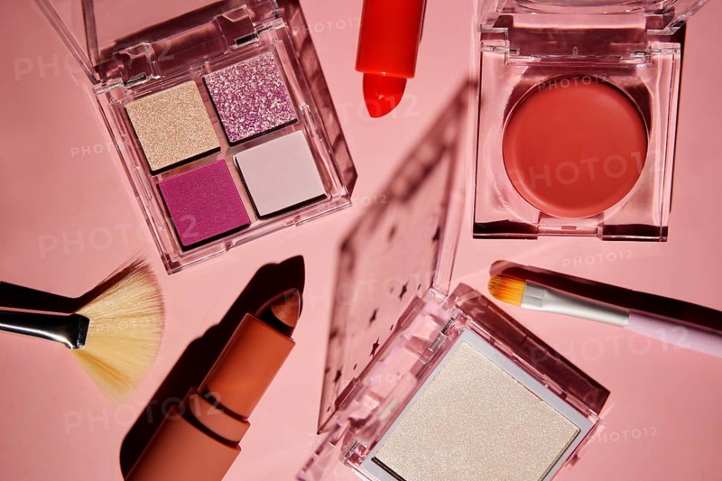 Top view of a vibrant makeup set on a pink background, featuring eyeshadow palettes, a lipstick, blush, brushes, and a creamy compact. The vivid colors enhance the pink tones
