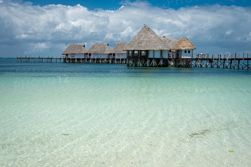 A beautiful row of traditional thatched-roof overwater bungalows connected by a wooden pier, extending over crystal-clear turquoise ocean water under a dramatic sky with fluffy white clouds in Zanzibar
