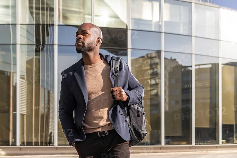 A professional cuban man in a blazer walks with confidence in a cityscape. The glass building behind reflects modern urban life, capturing dynamism and purpose