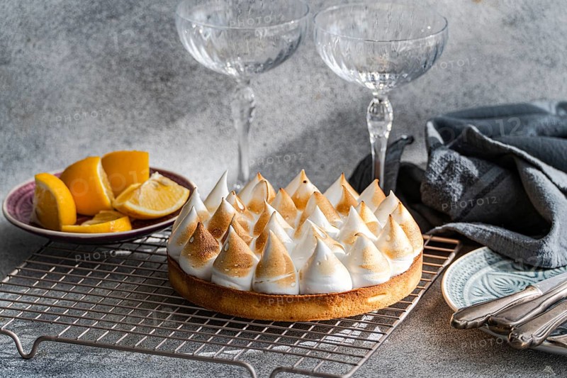A homemade lemon cake on a cooling rack beside sliced lemons. Two empty glasses add a touch of elegance, perfect for a dessert setting. Soft lighting enhances the scene