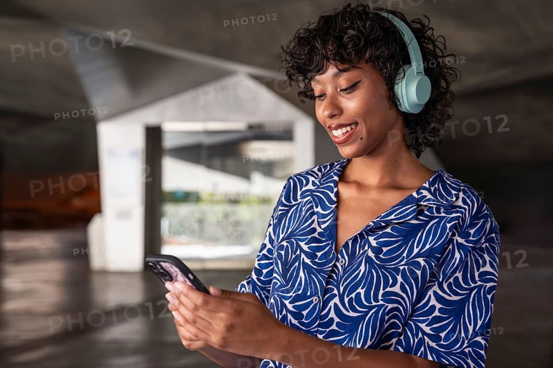A woman wearing blue headphones smiles while looking at her phone, standing in a modern architectural space. She wears a blue and white patterned shirt