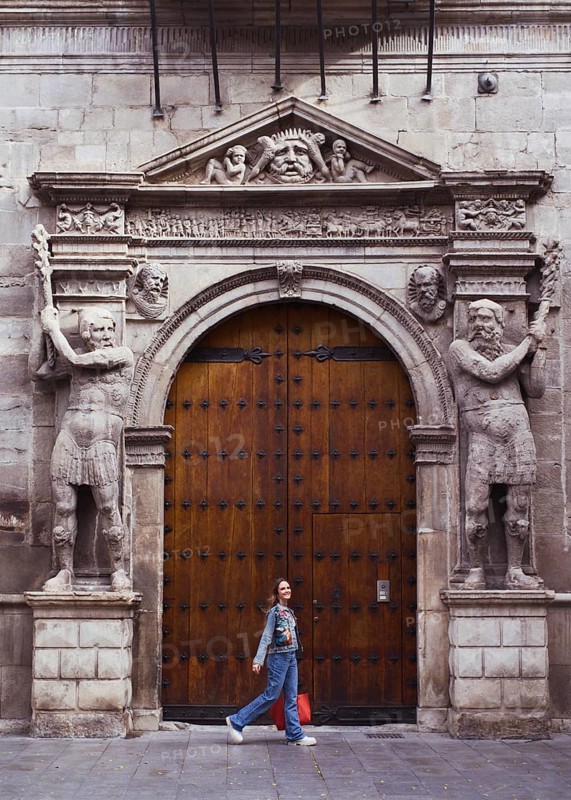 A young woman walks by an ornately carved wooden entrance, framed by sculpted stone figures. The majestic architecture hints at a historical building, blending past with present