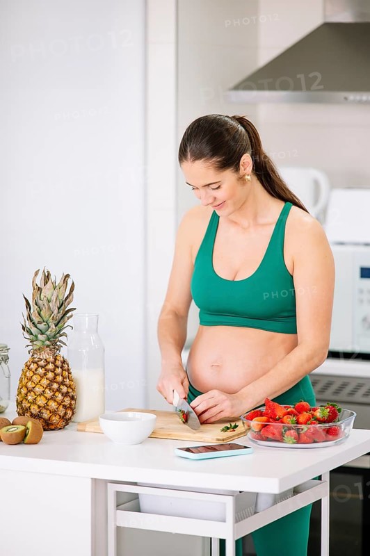 A pregnant woman in a green yoga outfit stands in a kitchen slicing strawberries. A variety of fresh fruits and a bottle of milk sit on the counter, emphasizing healthy eating