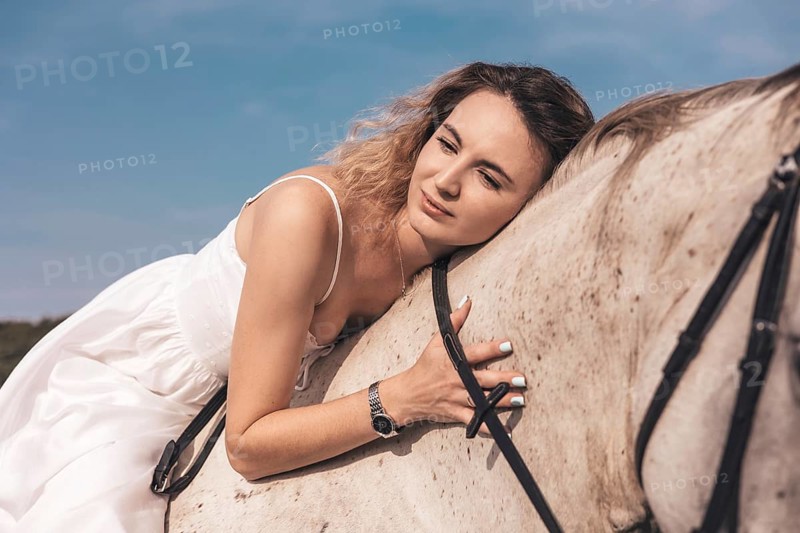 A young woman leans gently on a horse, enjoying a tranquil moment in a sunny field. Wearing a white dress, she exudes peace and connection with nature
