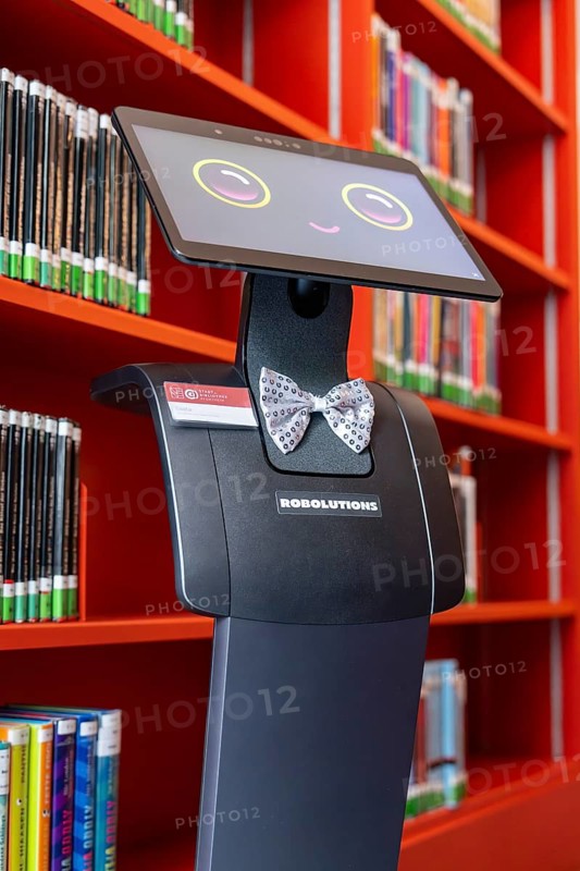 The robot stands in a library next to red bookshelves, interactive guided tours by robots Personalities from Robolutions, Pforzheim, Germany