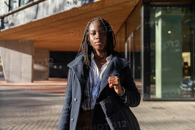 An African American businesswoman with braided hair enjoys a break outdoors, dressed smartly in a coat over a white shirt