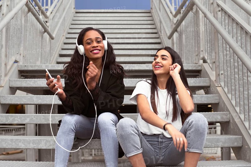Two young women sitting on outdoor stairs share a joyful moment while listening to music. One holds a phone and wears headphones, exuding happiness and connection