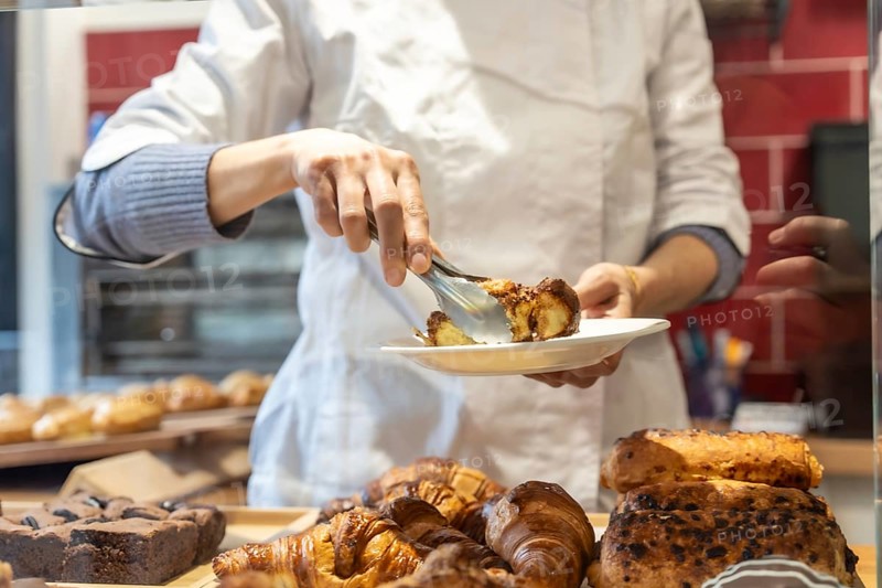 Baker wearing an apron and blue sweater serving a piece of chocolate pastry from a display case onto a white plate using metal tongs, with other baked goods visible