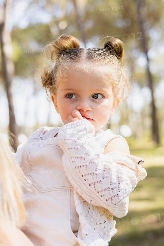 A young girl in her mother's arms gazes curiously at her surroundings during a sunny day outdoors. The scene captures a moment of warmth, love, and exploration