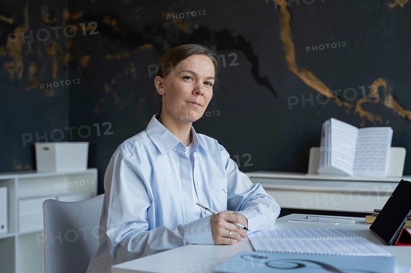 A focused woman sitting at a table writes notes for an upcoming piano lesson. She is surrounded by music sheets, in a calm setting, planning her instructional session