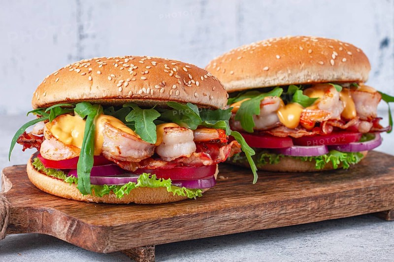 Two shrimp burgers and buns with sesame seeds, bacon, lettuce, tomatoes and onions, On a wooden board