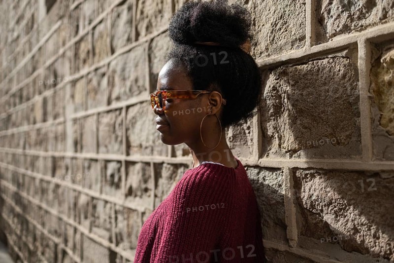A stylish woman in a burgundy sweater wearing sunglasses stands confidently against a textured stone wall, casting a shadow in the warm sunlight of a clear day