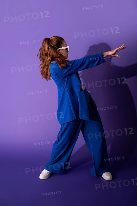A young woman dancer in a stylish blue outfit poses dynamically against a vibrant purple background. Her attire and confident stance convey energy and modernity