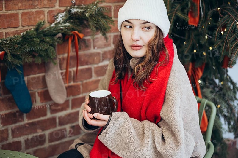 A young woman in a white beanie and red scarf holds a hot drink, seated outdoor on a snowy day, with festive decorations in the background