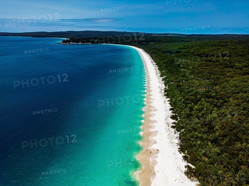 Aerial view of a pristine beach with white sand, turquoise waves, and lush greenery along the coastline under a clear blue sky, creating a serene and picturesque landscape in Jervis Bay, Australia