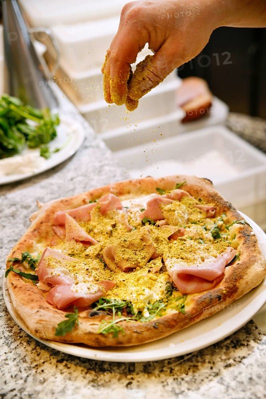Cropped unrecognizable person sprinkling seasoning on a homemade pizza topped with ham and arugula, in a kitchen setting