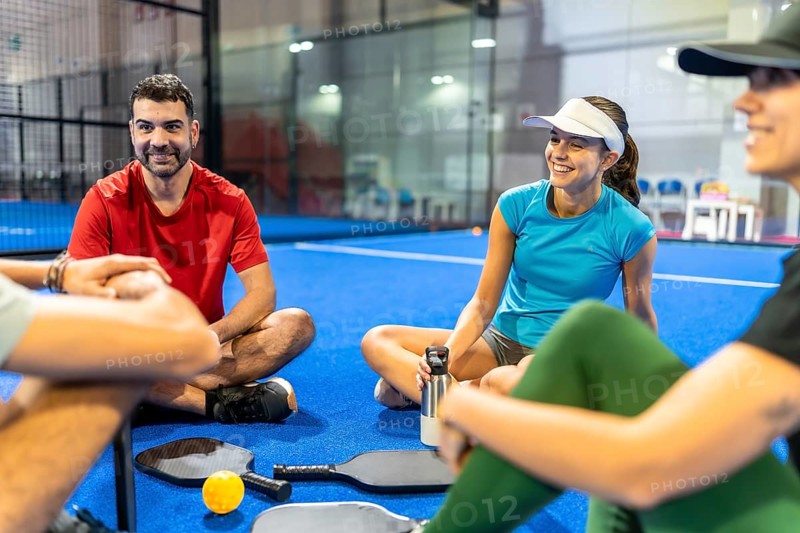 Group of young friends enjoying a break, sitting and smiling on the blue pickleball court with rackets and equipment after an active game, sharing happy moments and building friendship