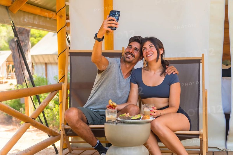 Happy couple taking a selfie with smartphone while enjoying a healthy breakfast on the porch of their glamping tent, celebrating their vacation and creating memories