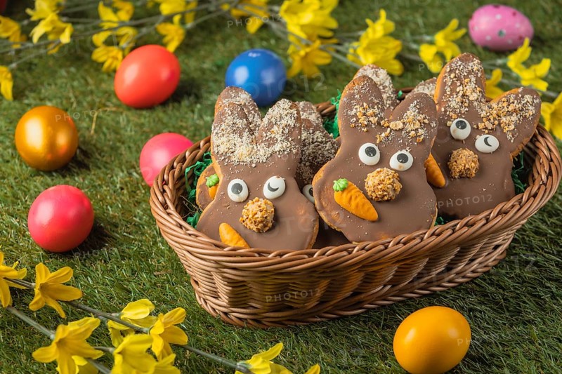 Bunny shaped Easter cookies with chocolate and carrot in a basket surrounded by colorful eggs on grass