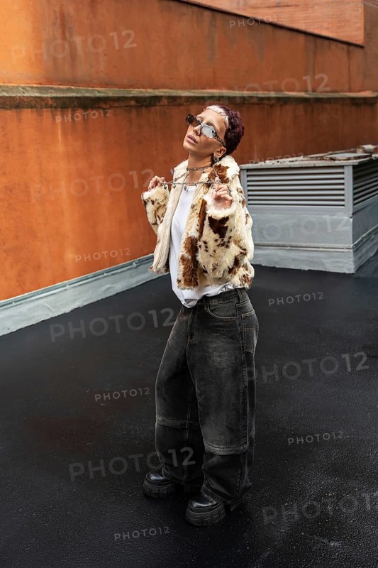 Fashionable woman in stylish faux fur jacket and sunglasses poses confidently against an urban background featuring gray and orange walls. The outdoor scene exudes modern elegance