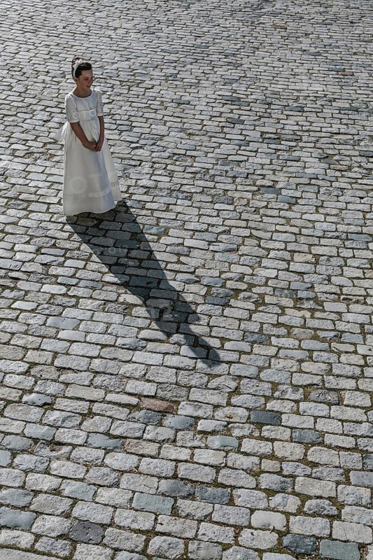 A young girl stands gracefully in a traditional First Communion dress on a cobblestone path, casting a long shadow. The scene captures the innocence and elegance of the occasion