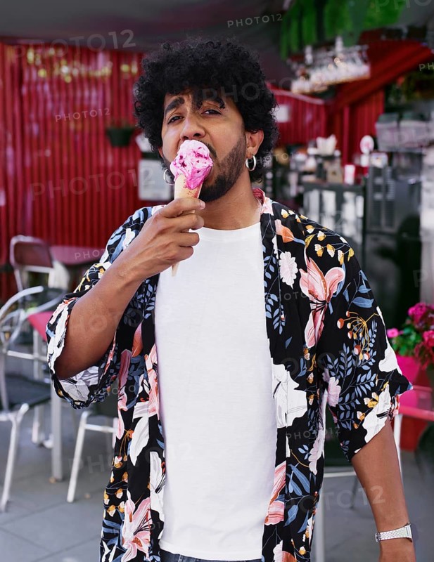 A man in a vibrant floral shirt enjoys a delicious ice cream cone during a sunny summer day. He stands near an outdoor cafe, savoring the treat with delight