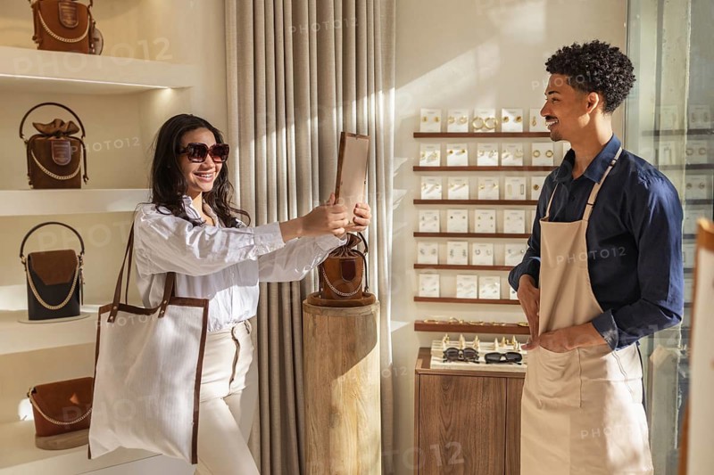 A cheerful woman shops in a stylish bag store, looking a mirror. Sunlight streams in, highlighting fashionable bags on display and creating a cozy atmosphere