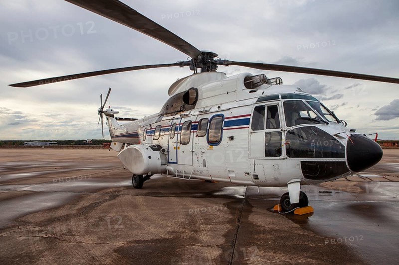 A white military helicopter with blue and red stripes is parked on the wet tarmac at an air base. The large rotor blades and sturdy structure highlight its transport capabilities