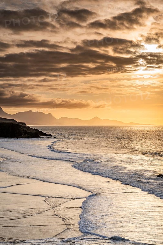 Gentle ocean waves wash onto the shore of La Pared Beach in Fuerteventura at sunset. The golden light and rugged mountains in the distance create a tranquil coastal atmosphere