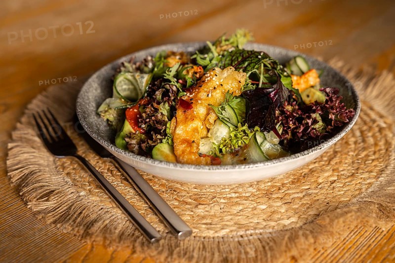 A vibrant green salad topped with crispy king prawns in batter, served in a ceramic bowl on a rustic wooden table with natural light enhancing its fresh, colorful appeal