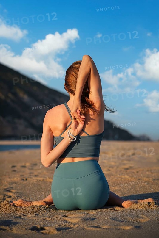 A woman sits in cow face pose at the beach, stretching her arms and enjoying the sunset by the water with serene clouds above