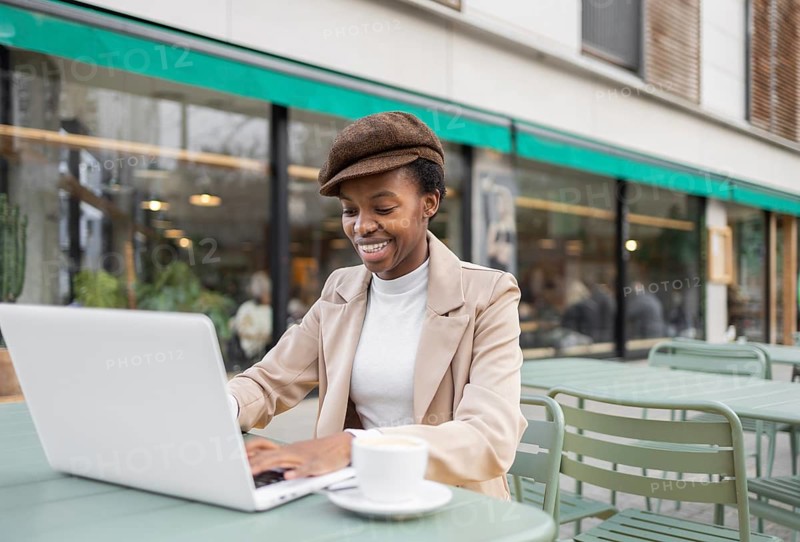 A black woman smiles while working on her laptop at an outdoor cafe Her stylish outfit and relaxed demeanor reflect a vibrant urban lifestyle and a blend of work and leisure