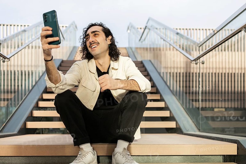 A young man with curly hair and a mustache wearing casual attire sits on outdoor steps, holding a smartphone and taking a selfie The environment is modern and urban