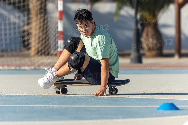 A teenager with Down Syndrome confidently skateboarding while sitting on a board, wearing protective gear, showcasing inclusivity, joy, and active participation in outdoor activities