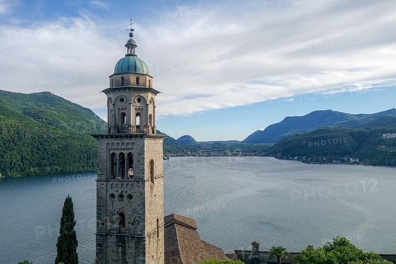 A picturesque scene at Lake Lugano in Ticino, Switzerland, featuring a majestic historic church tower with a backdrop of lush green mountains