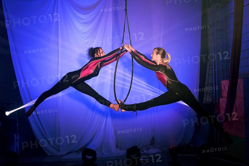 Two trapeze artists perform a captivating routine on a hoop, exhibiting grace and balance. Dressed in matching vibrant costumes, they are set against a dramatic blue lit background