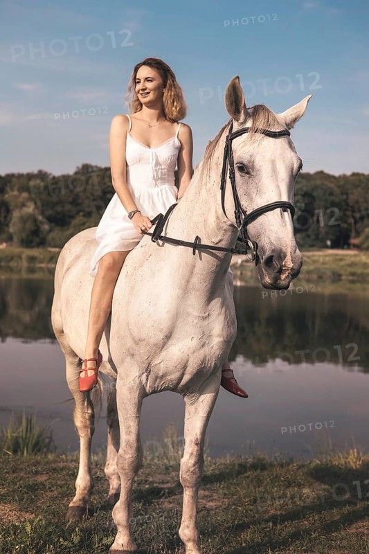 A young woman in a white dress is riding a white horse beside a calm lake. The serene landscape and sunny weather create a peaceful, idyllic scene. Perfect for themes of nature and leisure
