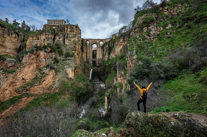 A traveler with a backpack standing on a rock with raised arms, overlooking the spectacular Puente Nuevo bridge and deep canyon in Ronda, Spain