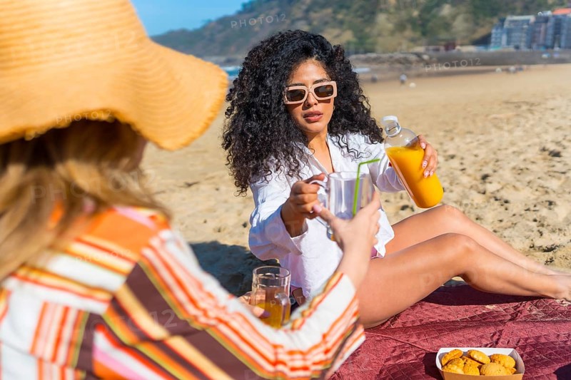 Two diverse women friends enjoying a summer picnic on a sandy beach, one pouring refreshing orange juice into a glass while relaxing under the clear sky