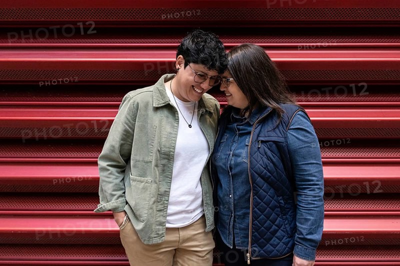 A curvy lesbian couple share a light heart moment, smiling warmly as they lean into each other. They're dressed casually, standing against a vibrant red backdrop, creating a joyful scene