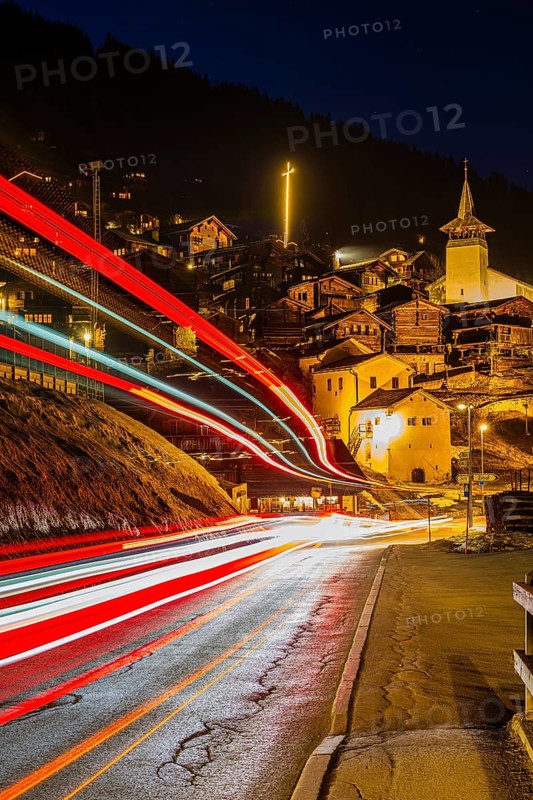 Braking track of a passing vehicle, old farmhouses and barn storage in the back, white cellars, the illuminated cross and the illuminated church tower of the Saint Theodule church, night view, historic town center, Grimentz, Val d'Anniviers, Valais Alps, Canton of Valais, Switzerland