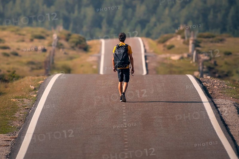 A solitary man with a backpack walks down a deserted winding road through verdant scenery in Guadarrama mountain range, Madrid. The image captures the essence of freedom, adventure, and exploration