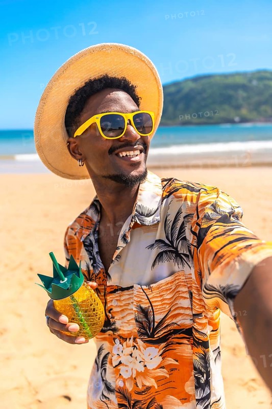 Young man wearing sunglasses and straw hat enjoys a tropical drink on a sunny beach while taking a selfie