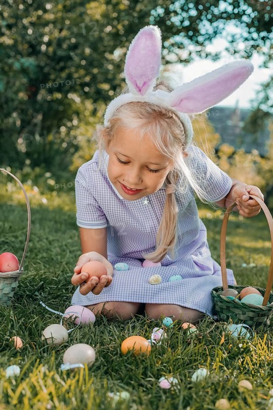 Young girl with bunny ears collects colorful Easter eggs in a sunny garden. She kneels on the grass, holding a basket, surrounded by eggs